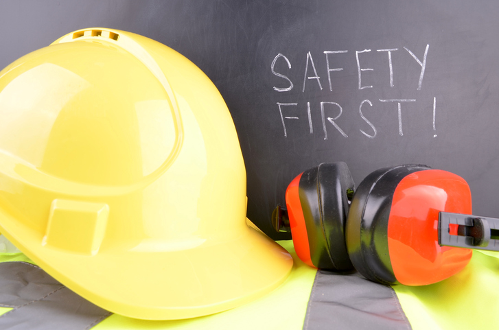 Safety first handwritten in white chalk with a yellow hard hat and red ear muffs in the fore ground.